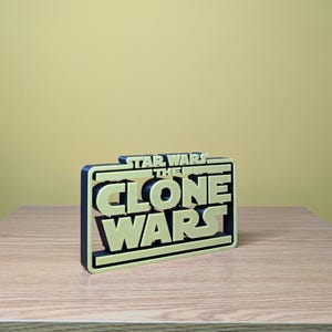 May include: A 3D printed, black and yellow sign with the text "Star Wars The Clone Wars" in a bold, blocky font.