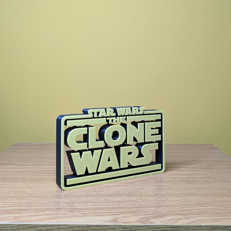 May include: A 3D printed, black and yellow sign with the text "Star Wars The Clone Wars" in a bold, blocky font.