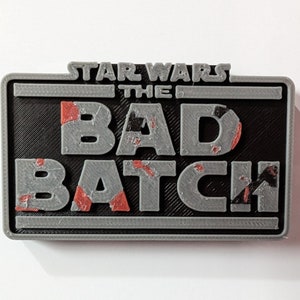 Op de afbeelding: Een grijs 3D-geprint bord met de tekst "STAR WARS THE BAD BATCH" in een verweerde stijl. Het bord heeft een zwarte achtergrond en rode en zwarte accenten.