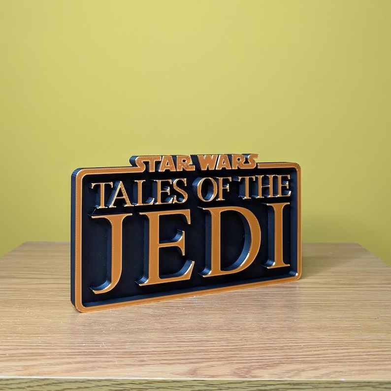 May include: A black and gold Star Wars "Tales of the Jedi" sign. The sign has a rectangular shape with the words "STAR WARS" at the top and "TALES OF THE JEDI" in large, bold letters. The sign is placed on a wooden surface.