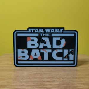 May include: A black and white sign with the text "Star Wars The Bad Batch" in white letters. The background of the sign is a light blue color with a camouflage pattern in orange.