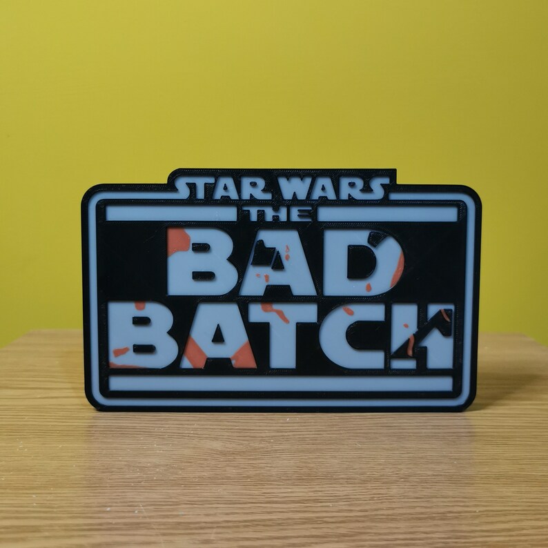 May include: A black and white sign with the text "Star Wars The Bad Batch" in white letters. The background of the sign is a light blue color with a camouflage pattern in orange.