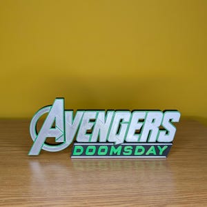 以下が含まれることがあります： 緑色の縁取りのある白字で「Avengers」と、黒いベースに緑字で「DOOMSDAY」と書かれた3Dプリントのサイン。 黄色の背景に対して、木製の表面に表示されています。