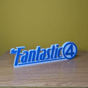 May include: A white and blue 3D-printed sign featuring the text "the Fantastic 4". The text is in a stylized font with a blue outline and a white interior. The number "4" is inside a blue circle.