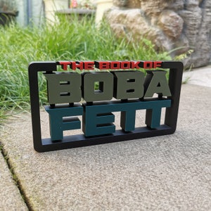 Peut inclure: Un cadre noir avec les mots "THE BOOK OF BOBA FETT" en lettres 3D. Les lettres sont vertes, bleues et rouges.