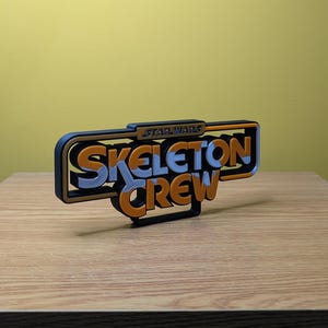 May include: A 3D printed sign with the text "STAR WARS SKELETON CREW" in orange and silver letters on a black background.