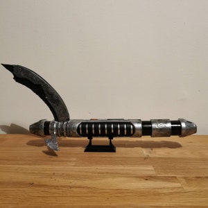 May include: A silver and black lightsaber with a curved hilt and a black stand. The lightsaber has a weathered, distressed look.