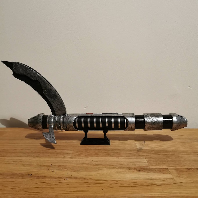 May include: A silver and black lightsaber with a curved hilt and a black stand. The lightsaber has a weathered, distressed look.