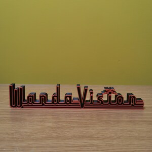 Peut inclure: Panneau imprim&eacute; en 3D noir et rouge avec l'inscription "WandaVision" dans une police stylis&eacute;e. Le panneau est pos&eacute; sur une surface en bois avec un fond jaune. Le design est de style r&eacute;tro.