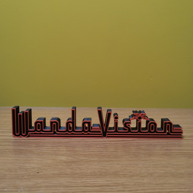 Peut inclure: Panneau imprim&eacute; en 3D noir et rouge avec l'inscription "WandaVision" dans une police stylis&eacute;e. Le panneau est pos&eacute; sur une surface en bois avec un fond jaune. Le design est de style r&eacute;tro.
