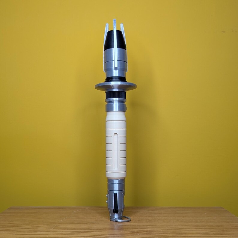 May include: A white and silver lightsaber with a black handle and a silver tip. The lightsaber is on a wooden surface.