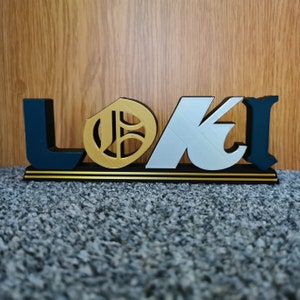 May include: A wooden sign with the word "LOKI" in black, gold, and silver letters. The "O" is a gold circle with a black design inside.