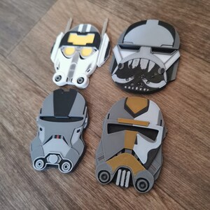 May include: Four Star Wars themed helmet figurines. The helmets are made of plastic and are painted in various shades of gray, white, and black. One helmet has a yellow stripe and a yellow visor. Another helmet has a gold stripe and a gold visor. The helmets are all about 2 inches tall.