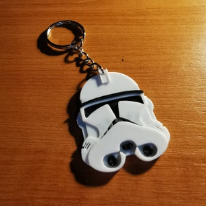 May include: White plastic keychain shaped like a Star Wars Stormtrooper helmet. The helmet has a black visor and a black stripe across the top.