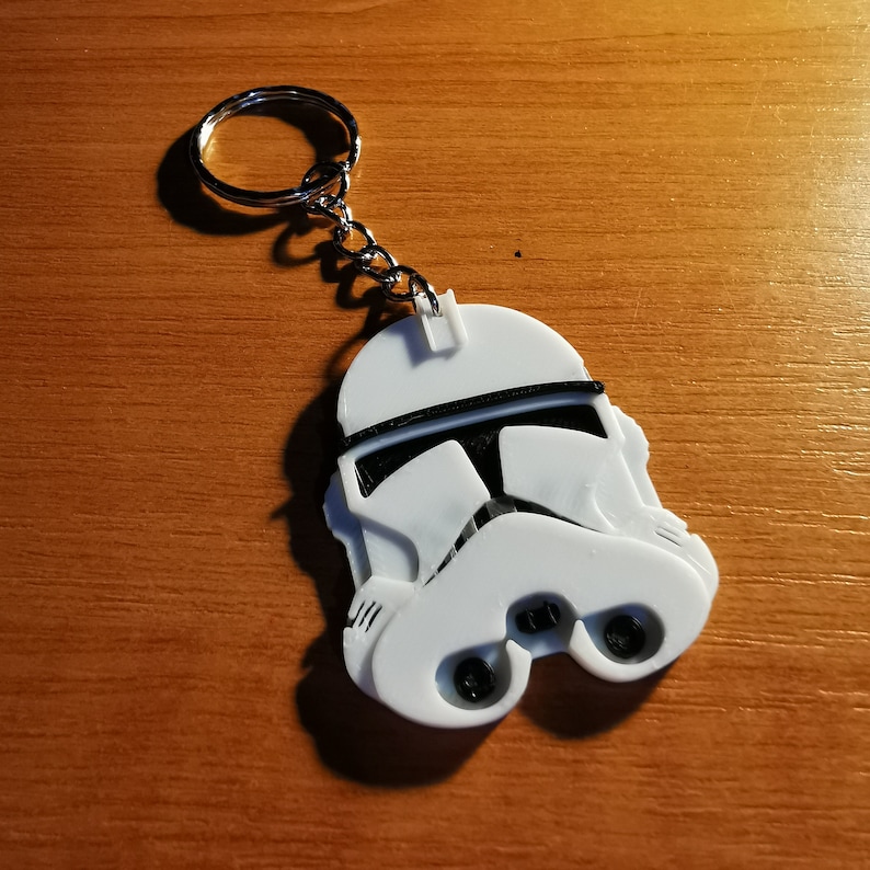 May include: White plastic keychain shaped like a Star Wars Stormtrooper helmet. The helmet has a black visor and a black stripe across the top.