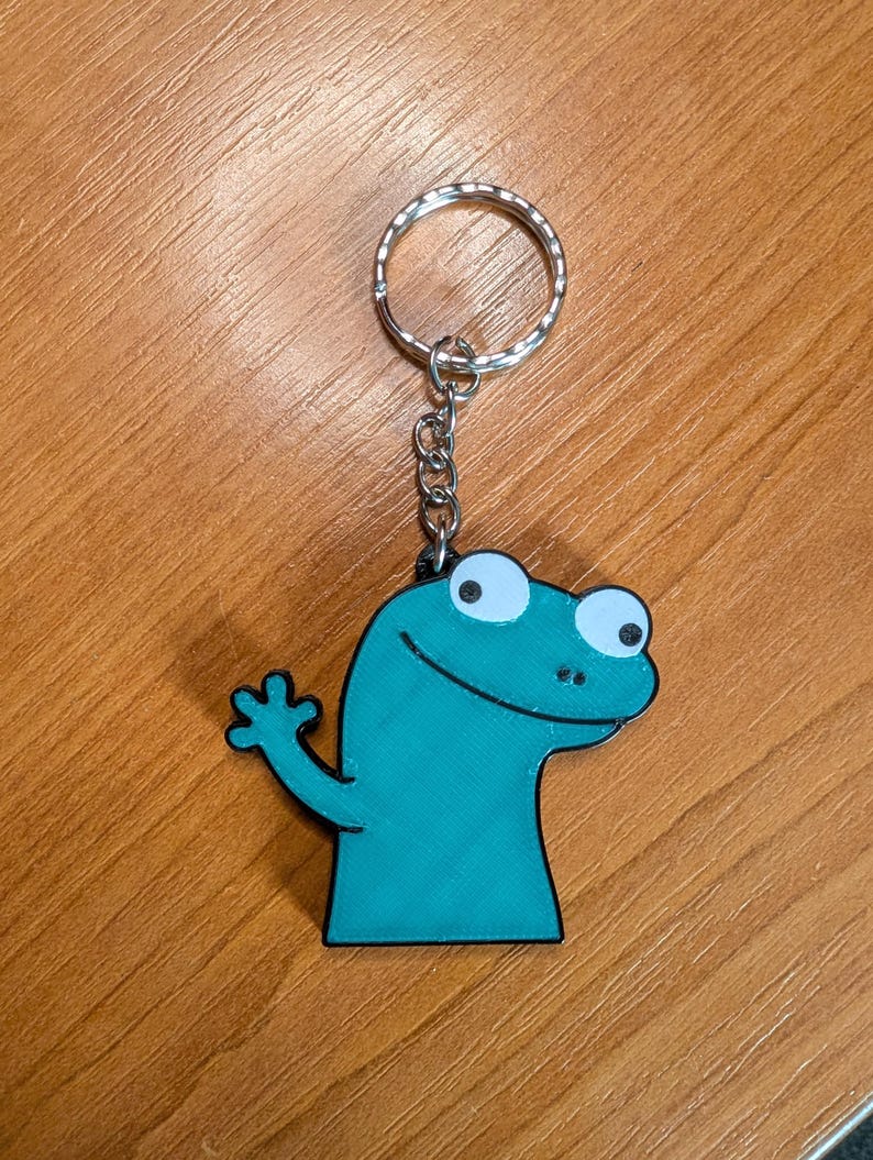 Peut inclure: Un porte-cl&eacute;s en forme de l&eacute;zard de dessin anim&eacute; turquoise avec un contour noir. Le l&eacute;zard a des yeux blancs et l&egrave;ve la main droite. Le porte-cl&eacute;s a un anneau et une cha&icirc;ne argent&eacute;s.
