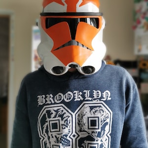May include: A person wearing an orange and white Star Wars clone trooper helmet with a black visor. The helmet has a white stripe across the top and a black stripe across the bottom. The person is wearing a blue hoodie with the word "BROOKLYN" printed on the front.