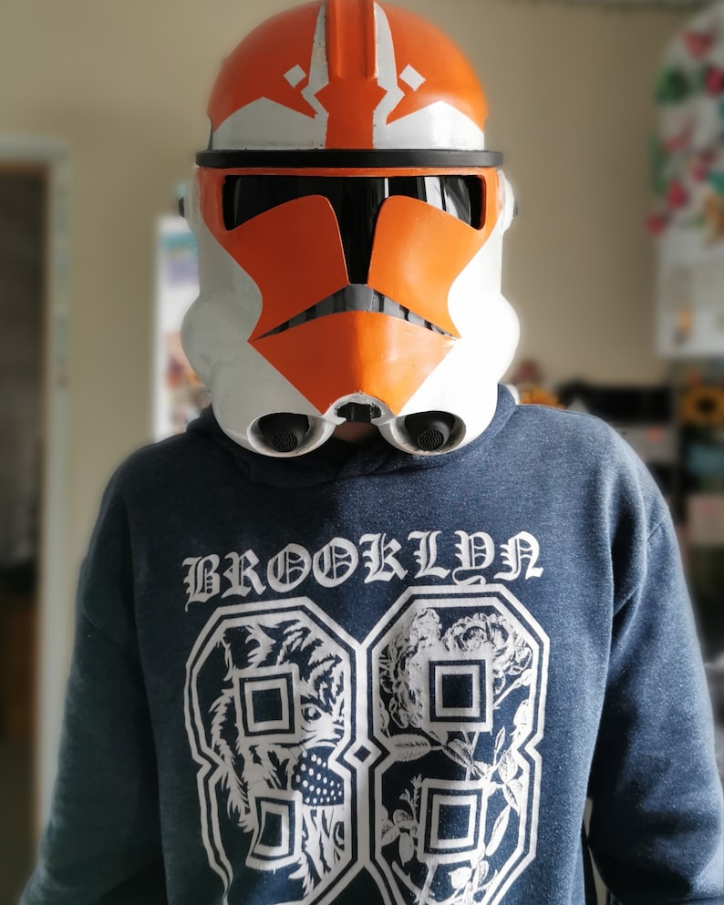 May include: A person wearing an orange and white Star Wars clone trooper helmet with a black visor. The helmet has a white stripe across the top and a black stripe across the bottom. The person is wearing a blue hoodie with the word "BROOKLYN" printed on the front.