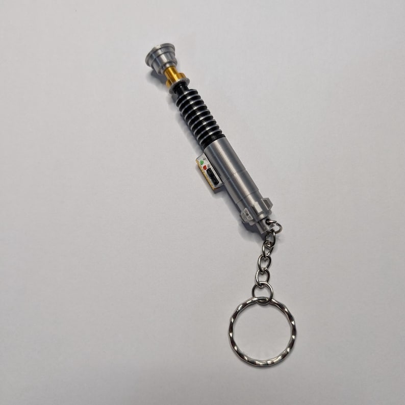 May include: A silver lightsaber keychain with black and gold accents. The keychain features a detailed replica of a lightsaber with a silver ring for attaching keys. The lightsaber has a black ribbed section and a small control panel.