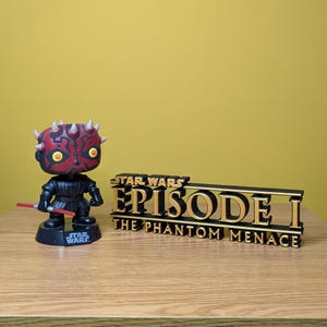 May include: A red and black Darth Maul Star Wars Pop figure stands in front of a black and gold Star Wars Episode I: The Phantom Menace logo.