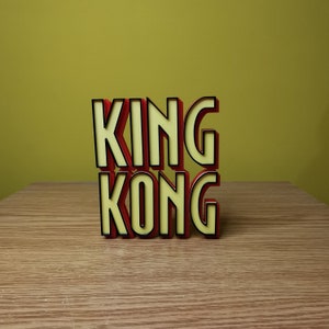 Pu&ograve; includere: Lettere 3D rosse e gialle che compongono la scritta "KING KONG" su una superficie di legno.