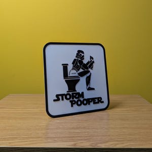 May include: White sign with a black border and a black and white illustration of a Stormtrooper sitting on a toilet. The text "Storm Pooper" is printed below the illustration.