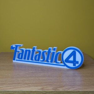 May include: A white and blue 3D-printed sign featuring the text "the Fantastic 4". The sign has a blue outline and a blue circle with the number 4. The sign is on a wooden surface with a yellow background.
