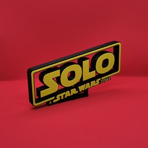 May include: A black and yellow 3D sign with the word "SOLO" in yellow letters and the text "A STAR WARS STORY" in black letters.