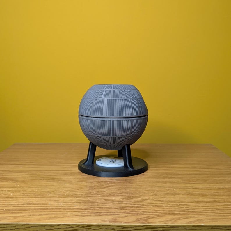 May include: A gray, spherical Death Star model with a textured surface, mounted on a black base with three legs.