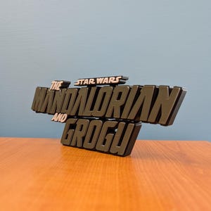 Op de afbeelding: Een 3D-bord met de tekst "THE MANDALORIAN AND GROGU" en "STAR WARS" erboven. Het bord is donkergroen en zwart, met een gelaagd ontwerp. Het staat op een houten oppervlak tegen een blauwe muur.