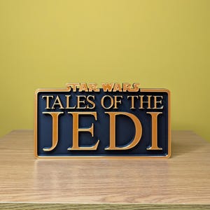 May include: A rectangular sign with the words "STAR WARS TALES OF THE JEDI" in gold lettering on a navy blue background. The sign is framed in gold and sits on a wooden surface.