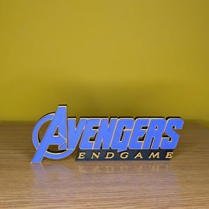 May include: A blue and gold "Avengers: Endgame" logo, standing upright on a wooden surface. The logo features the word "Avengers" in blue with gold trim, and "Endgame" in gold lettering.