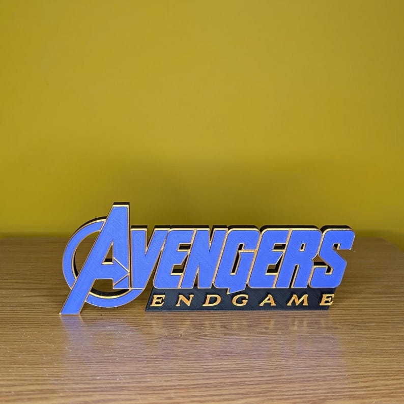 May include: A blue and gold "Avengers: Endgame" logo, standing upright on a wooden surface. The logo features the word "Avengers" in blue with gold trim, and "Endgame" in gold lettering.