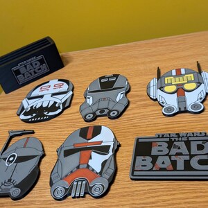 May include: Six Star Wars: The Bad Batch character enamel pins. The pins are in the shape of the characters' helmets. The pins are gray, white, and red. The pins are on a wooden surface. The text "Star Wars The Bad Batch" is on a black and gray enamel pin.