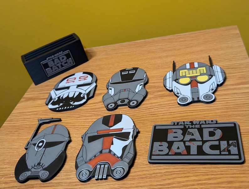 May include: Six Star Wars: The Bad Batch character enamel pins. The pins are in the shape of the characters' helmets. The pins are gray, white, and red. The pins are on a wooden surface. The text "Star Wars The Bad Batch" is on a black and gray enamel pin.