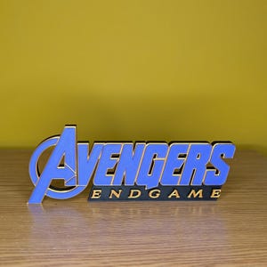 May include: A blue and gold Avengers Endgame logo, a 3D printed decorative item. The word "Avengers" is in blue with a gold outline, and "Endgame" is in gold lettering on a black base. The background is a solid yellow.