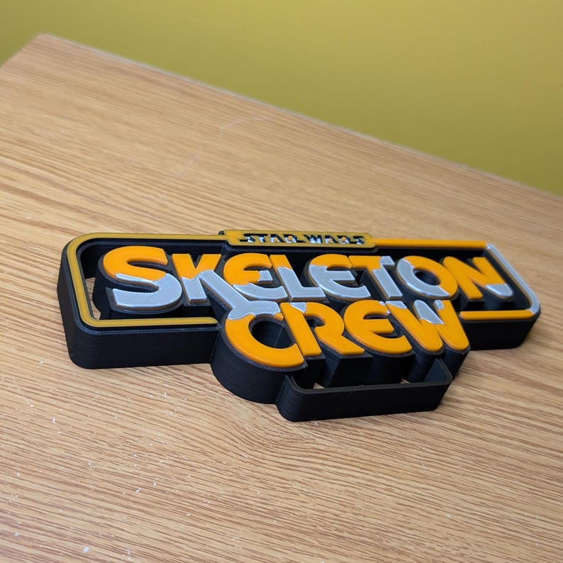 May include: A 3D printed black, yellow, and silver sign that reads "Star Wars Skeleton Crew".