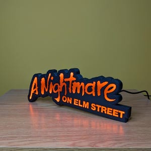 May include: Black and orange LED sign that reads "A Nightmare on Elm Street." The sign is shaped like a bat and sits on a wooden surface. The text is in a stylized font and glows orange.