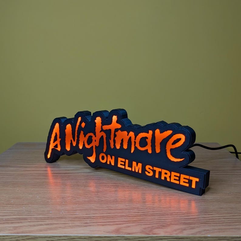 May include: Black and orange LED sign that reads "A Nightmare on Elm Street." The sign is shaped like a bat and sits on a wooden surface. The text is in a stylized font and glows orange.