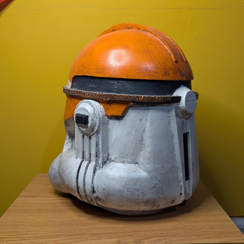 May include: A white and orange plastic helmet with a weathered look. The helmet has a black visor and a raised design on the front.