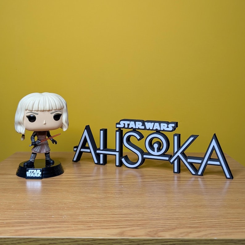 May include: A black and white Star Wars Ahsoka Tano logo with a white outline. A Funko Pop! figure of Ahsoka Tano is standing in front of the logo. The figure is wearing a brown and white outfit and holding a red lightsaber.