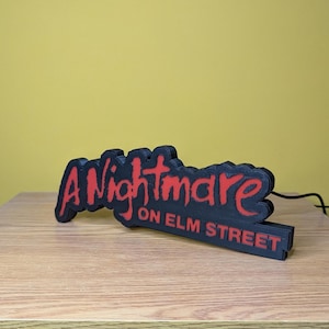 May include: A black and red sign that reads "A Nightmare on Elm Street." The sign is made of a dark material with the text in a bold, red font. The sign is on a wooden surface with a yellow background.