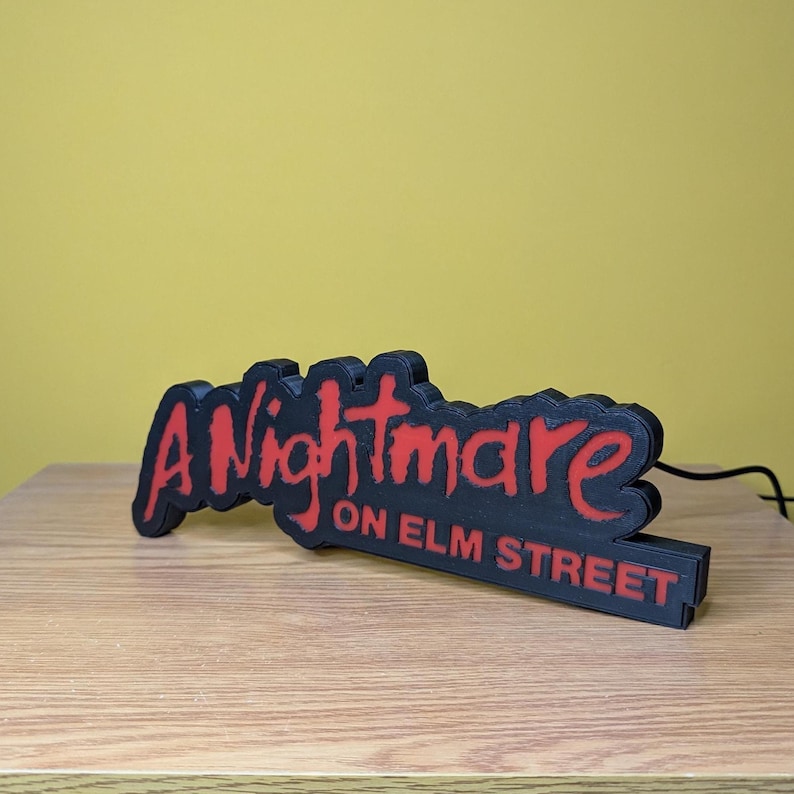 May include: A black and red sign that reads "A Nightmare on Elm Street." The sign is made of a dark material with the text in a bold, red font. The sign is on a wooden surface with a yellow background.