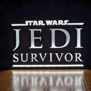 以下が含まれることがあります： 黒い文字で「STAR WARS JEDI SURVIVOR」と書かれた白い照明付き看板