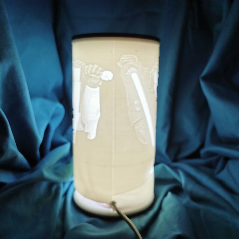 May include: White ceramic lamp with a 3D design of a person holding a lightsaber. The lamp is on a blue surface.