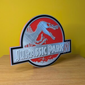 Op de afbeelding: 3D-geprint Jurassic Park-logo. Het heeft een rode achtergrond met een zilveren dinosaurus silhouet en de woorden "JURASSIC PARK" in het wit. Het logo heeft een zwarte rand en staat tegen een gele achtergrond.