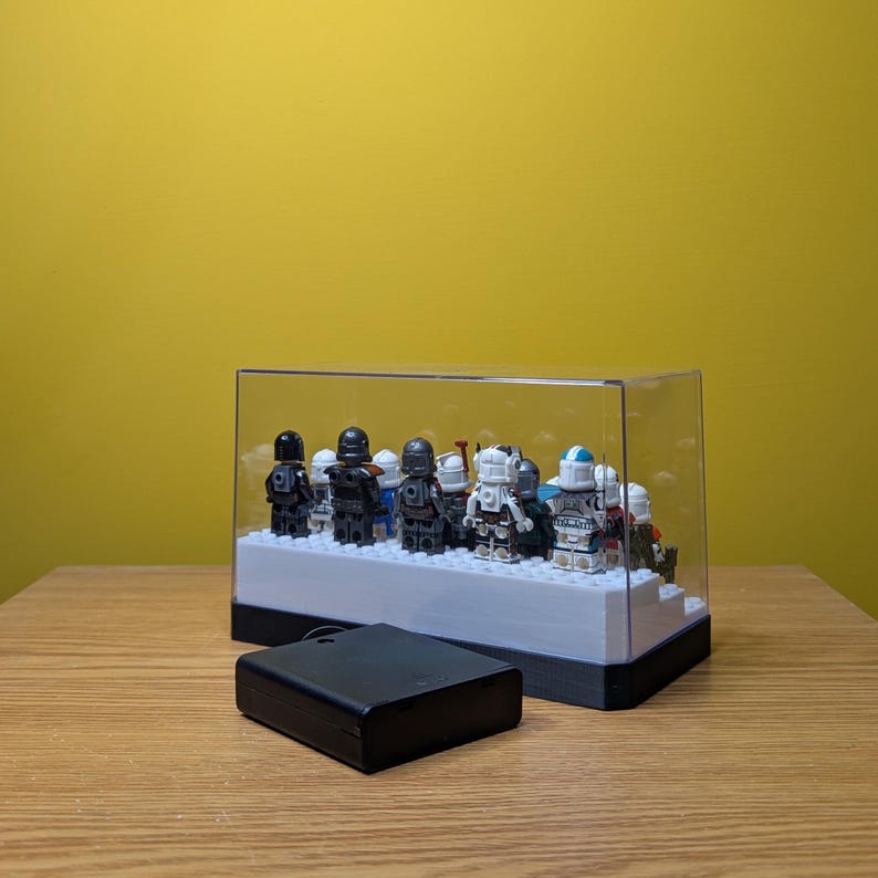 May include: A clear plastic display case with a black base, containing a collection of small, detailed figurines. The figurines are mostly black and gray, with some white and blue accents. The case is lit from below, illuminating the figurines.
