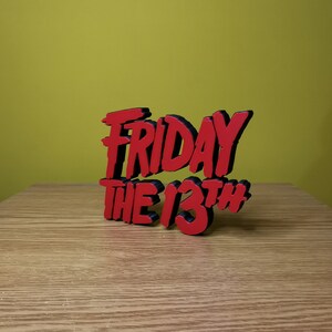 May include: A red and black 3D sign that reads "Friday the 13th" in a stylized font. The sign is sitting on a wooden surface.