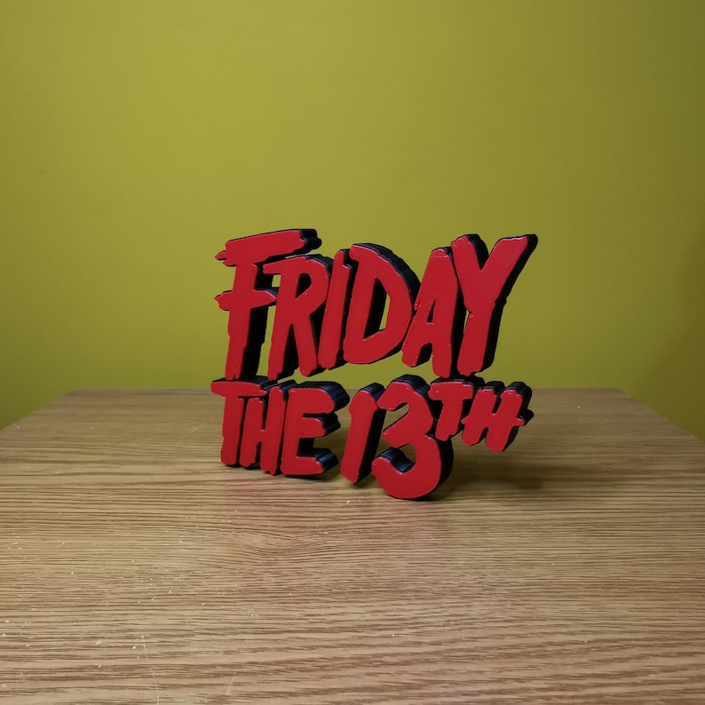 May include: A red and black 3D sign that reads "Friday the 13th" in a stylized font. The sign is sitting on a wooden surface.