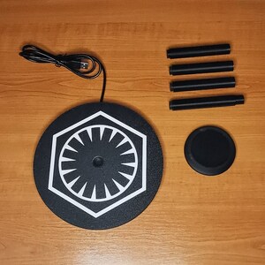 May include: A black circular charging stand with a white symbol resembling a star with multiple points. The stand is surrounded by four black metal rods, which are likely used for support or attachment. The stand is on a wooden surface.